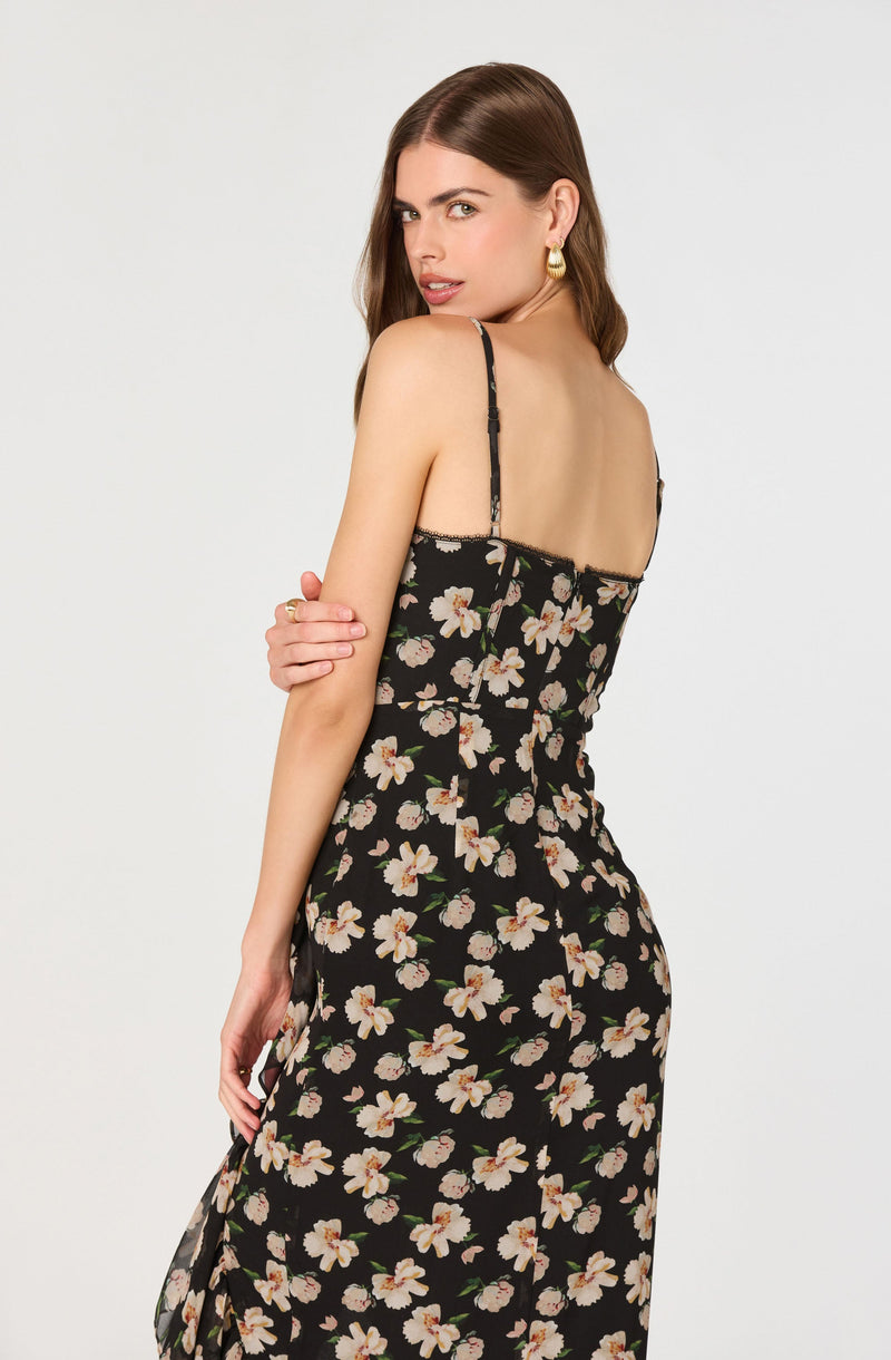 A woman is wearing a black dress with a floral pattern of white and pink flowers with green leaves. The dress has thin, adjustable spaghetti straps.