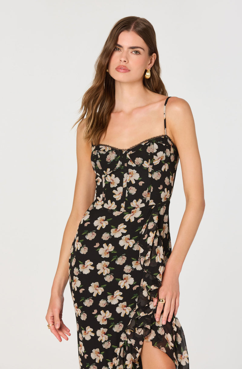 A woman is wearing a black dress with a floral pattern of large white and pink flowers with green leaves. The dress has thin spaghetti straps, a fitted bodice, and a ruffled detail running down the front. It appears to be made of a lightweight, flowy fabric.