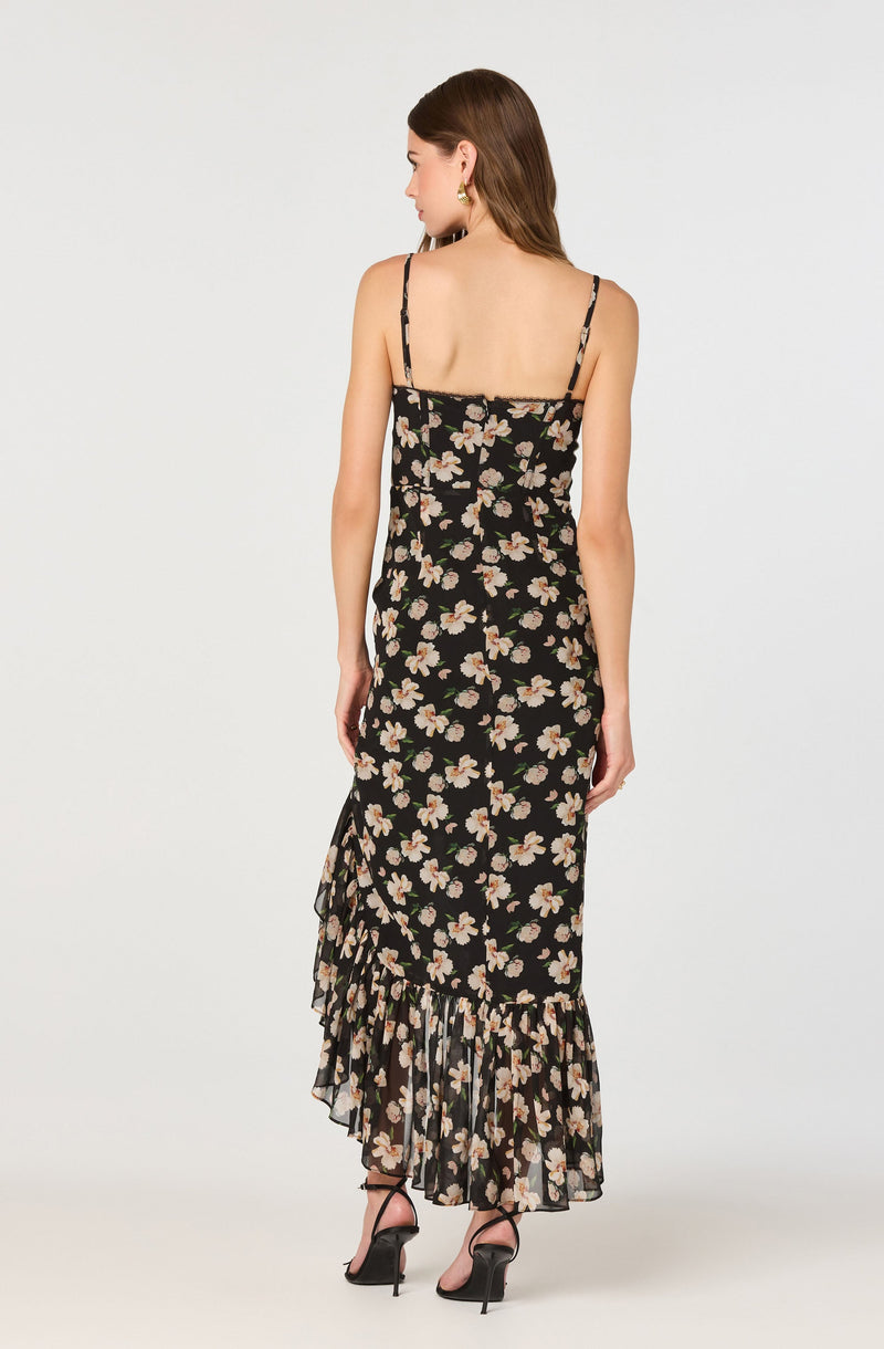 A woman is wearing a sleeveless, full-length dress with thin spaghetti straps. The dress features a black background with an all-over floral pattern of white and light pink flowers with green leaves. It has a ruffled hemline that adds a decorative touch at the bottom.