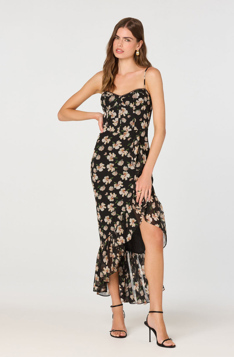A woman is wearing a sleeveless black dress with a floral pattern of light pink and beige flowers, thin spaghetti straps, and a midi length with ruffle details and a front slit. She is also wearing black high-heeled sandals with ankle straps.