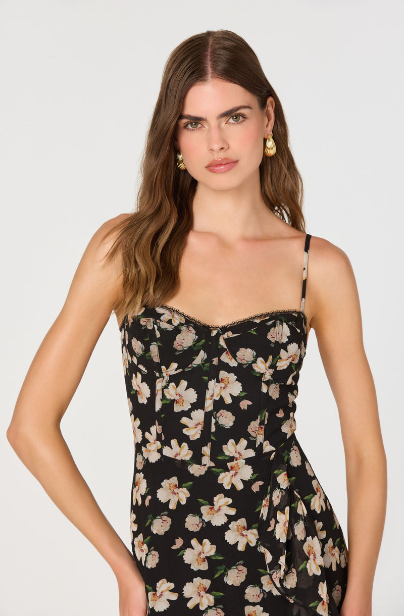 A woman is wearing a black dress with a floral pattern of white and pink flowers with green leaves. The dress has thin spaghetti straps and a fitted bodice. It features delicate black lace trim along the neckline and vertical seam details down the front.