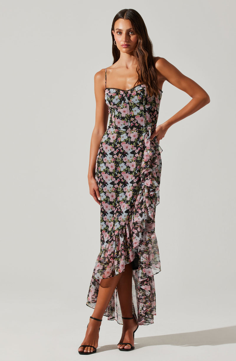 The dress is a floor-length, sleeveless design with thin spaghetti straps. It features a black background adorned with a floral print in shades of pink, lavender, and green. The dress has a fitted bodice, and down the side, there is a ruffled asymmetrical overlay that extends from the waist to the hem, creating a layered look. The fabric appears lightweight and semi-sheer, with a slit that reveals the legs underneath. The model pairs the dress with high-heeled sandals.