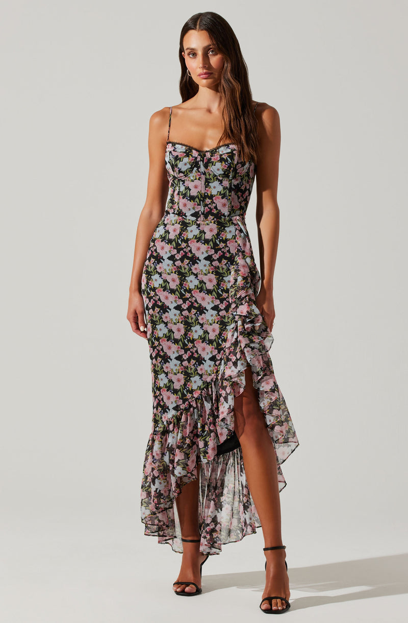 A woman is wearing a sleeveless, floral print dress with thin spaghetti straps and a ruffled asymmetric hemline. The dress features a black background with pink, white, and purple flowers and green leaves. It has a fitted bodice and a high-low ruffled skirt that is longer at the back. She is also wearing black high-heeled sandals.