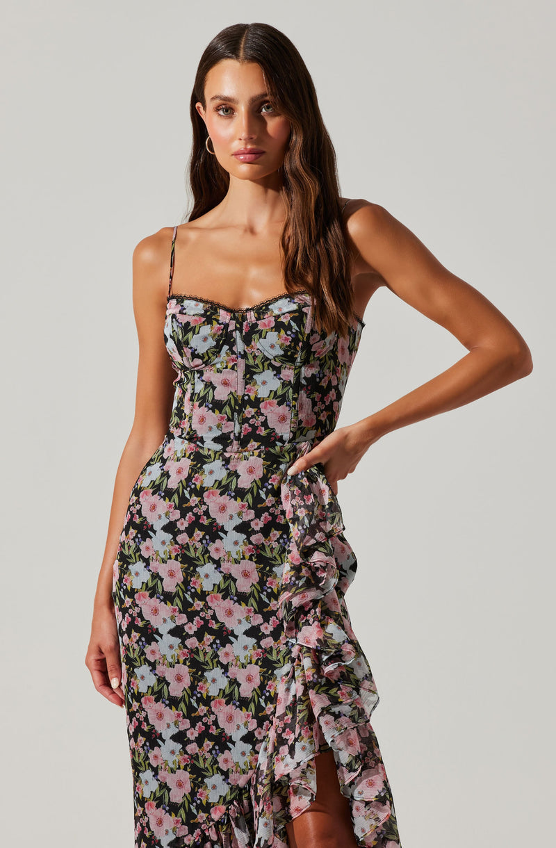 A woman is wearing a slip dress with thin spaghetti straps and a floral pattern featuring pink, white, and purple flowers with green leaves on a black background. The dress has a ruffled detail running along the side and down the front.