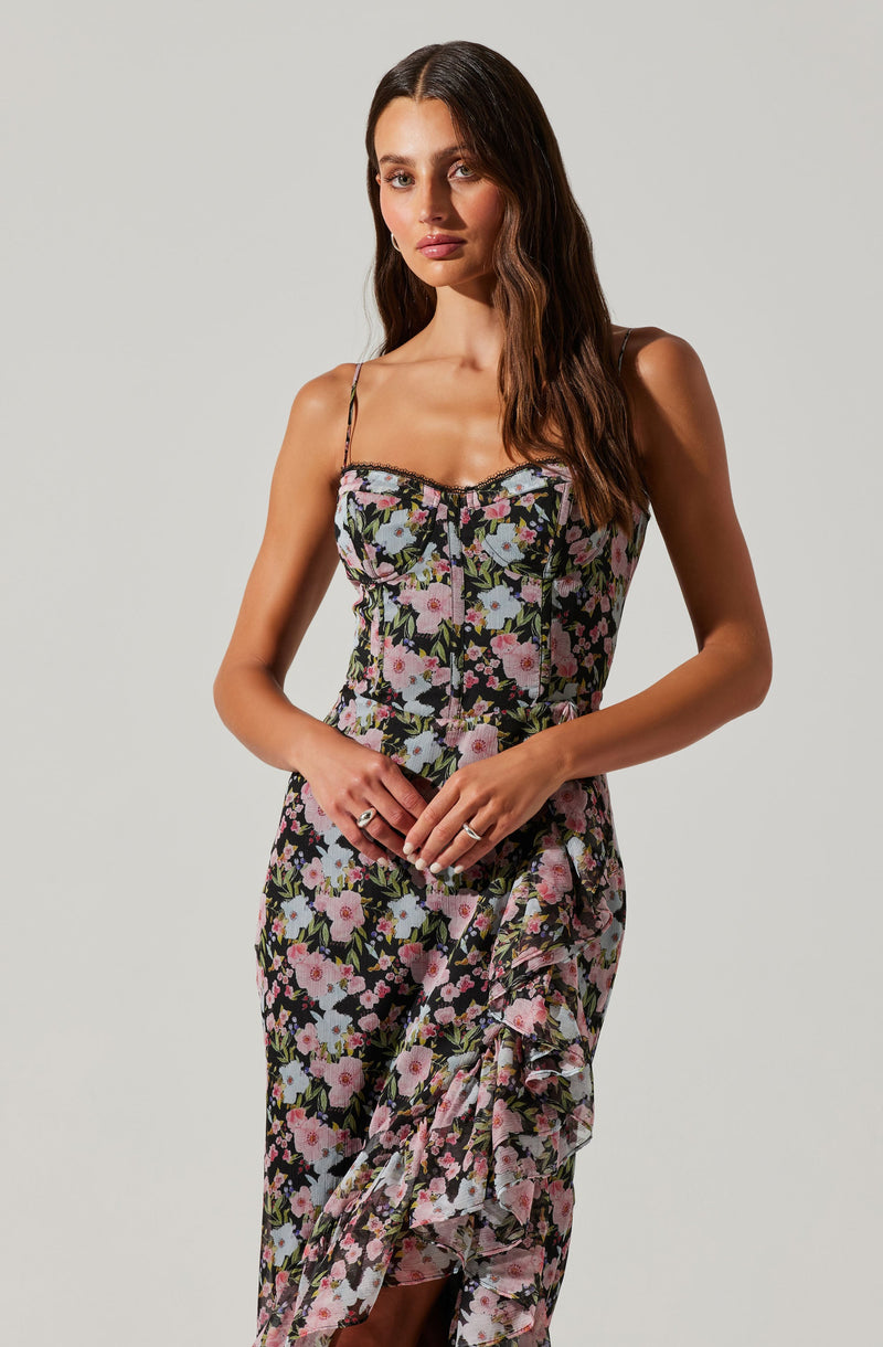 A woman is wearing a floral dress with a black background and pink, white, and light blue flowers. The dress has thin spaghetti straps and a fitted bodice. The skirt features a ruffle detail along one side that cascades down the length of the dress.
