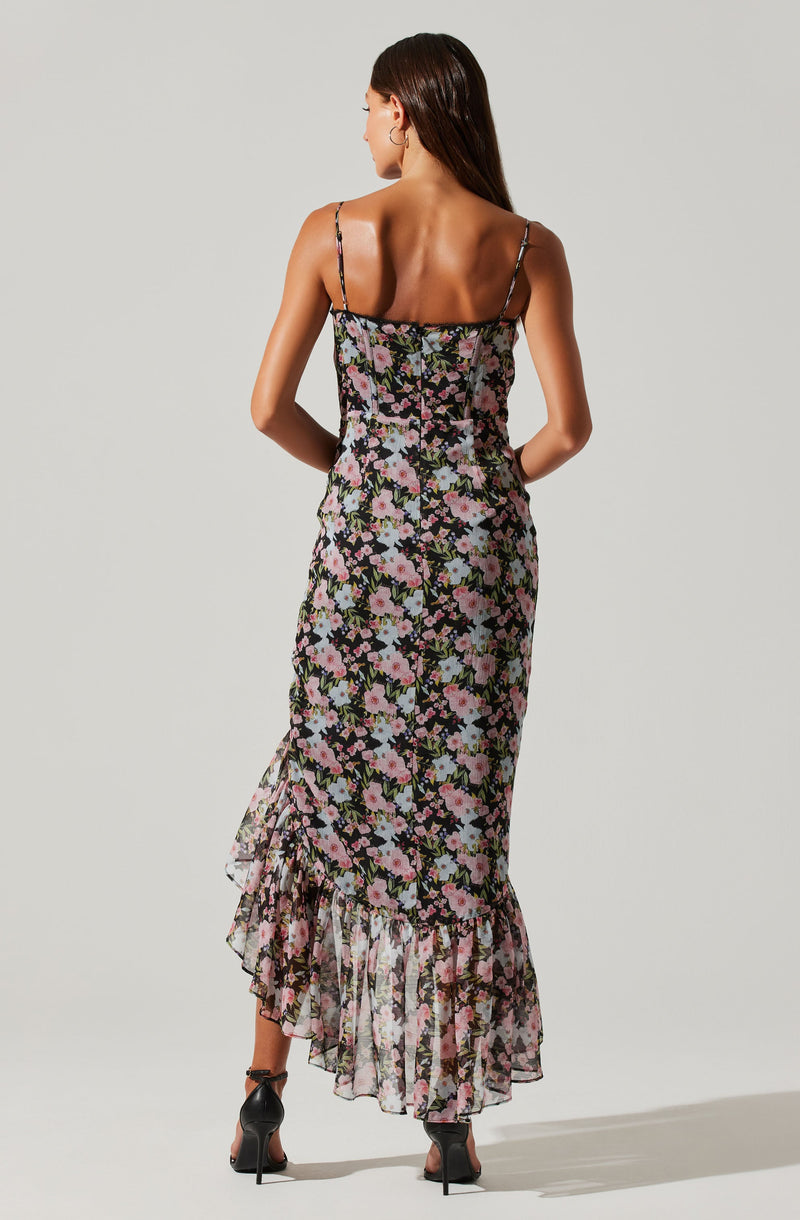 A woman is wearing a long, sleeveless dress with thin spaghetti straps. The dress features a floral pattern with pink, white, and purple flowers on a black background. The dress has a fitted bodice and a ruffled hemline that reaches the ankles. She is also wearing black high-heeled shoes.