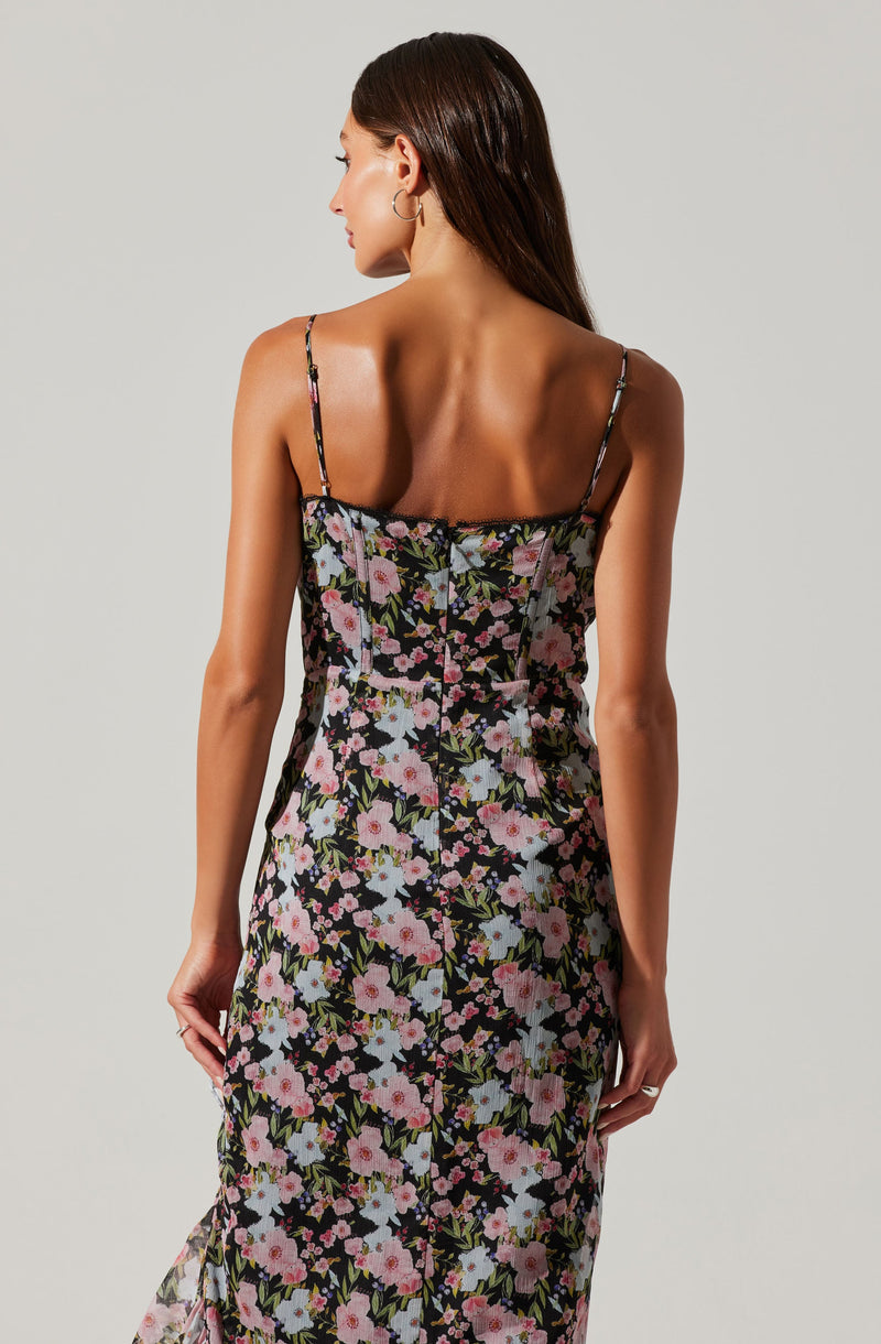 A woman is wearing a sleeveless, spaghetti strap dress with a floral pattern featuring pink, white, and purple flowers, along with green leaves on a black background.