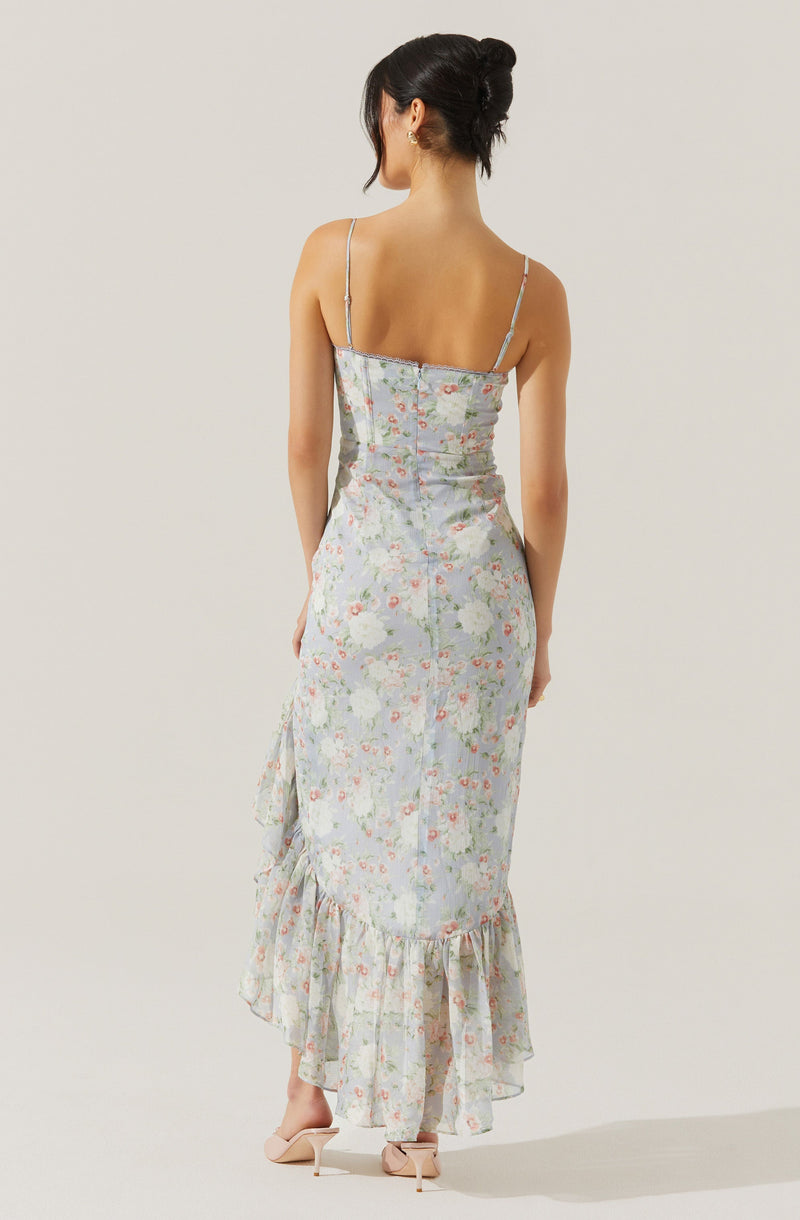 A woman is wearing a light-colored, floral-patterned dress with a fitted bodice and thin spaghetti straps. The dress has a ruffled hem that falls to ankle length and features a delicate, pastel floral print. She is also wearing nude high-heeled sandals.