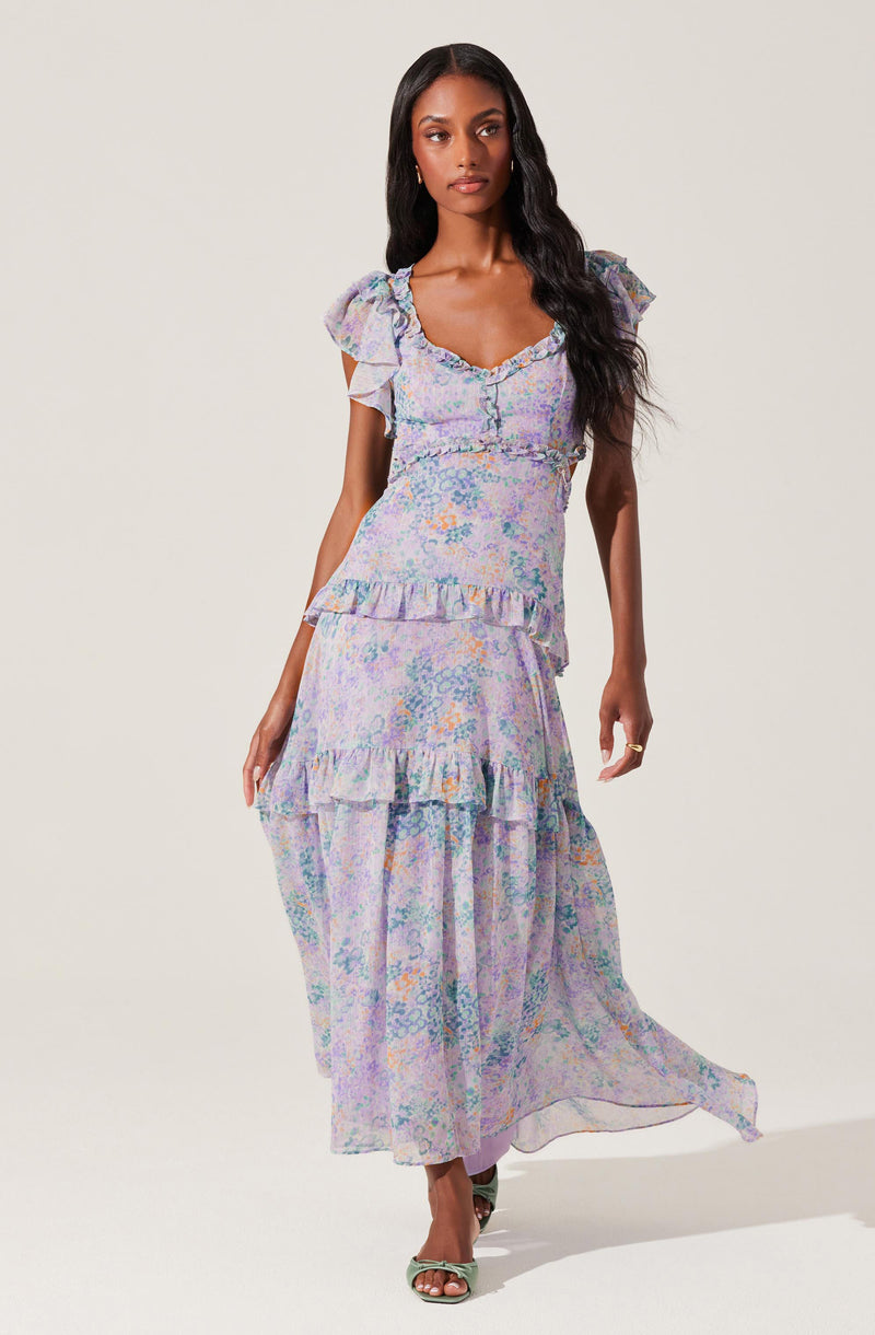 A woman is wearing a long, flowing dress with a pastel floral print in shades of purple, blue, and orange. The dress features short, ruffled sleeves, a fitted bodice with a sweetheart neckline, and ruffle details along the hem and tiers of the skirt.