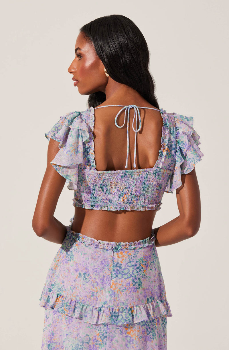 A woman is wearing a coordinated outfit featuring a cropped top and a skirt. The top has a square neckline with ruffled, cold-shoulder sleeves and is tied at the back with a thin string. The skirt has a high waist and a tiered, ruffled hem. The fabric of both pieces has a colorful, abstract floral pattern in shades of purple, blue, green, orange, and pink.