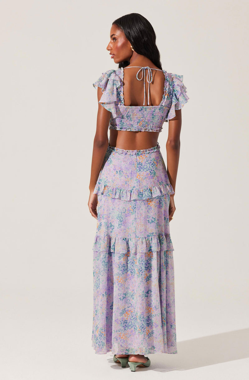 A woman is wearing a two-piece outfit made of a pastel-colored, floral-patterned fabric. The top is a cropped blouse with flutter sleeves and a square neckline, tied at the back with a bow. The matching skirt is high-waisted, with a tiered ruffle detail around the hips and a long, flowing length reaching the floor.