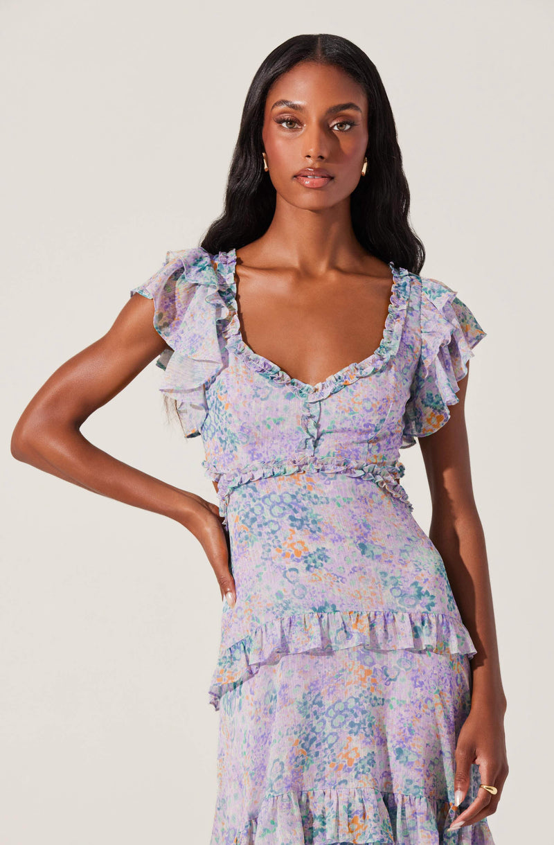 A woman is wearing a light-colored dress with a pastel, watercolor-like pattern that includes shades of lavender, orange, blue, and green. The dress features ruffled sleeves, ruffles around the neckline, and ruffles at the waist and hemline. The fabric appears lightweight and textured, with a fitted waist and a flowing skirt.