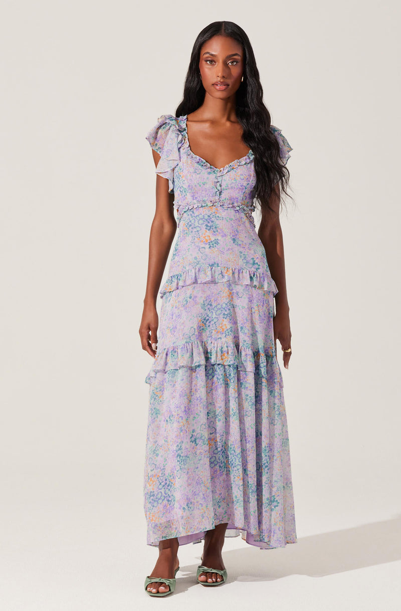 A woman is wearing a long, flowy dress with a pastel, colorful floral pattern. The dress features ruffled cap sleeves, a sweetheart neckline, and tiered ruffles along the skirt.