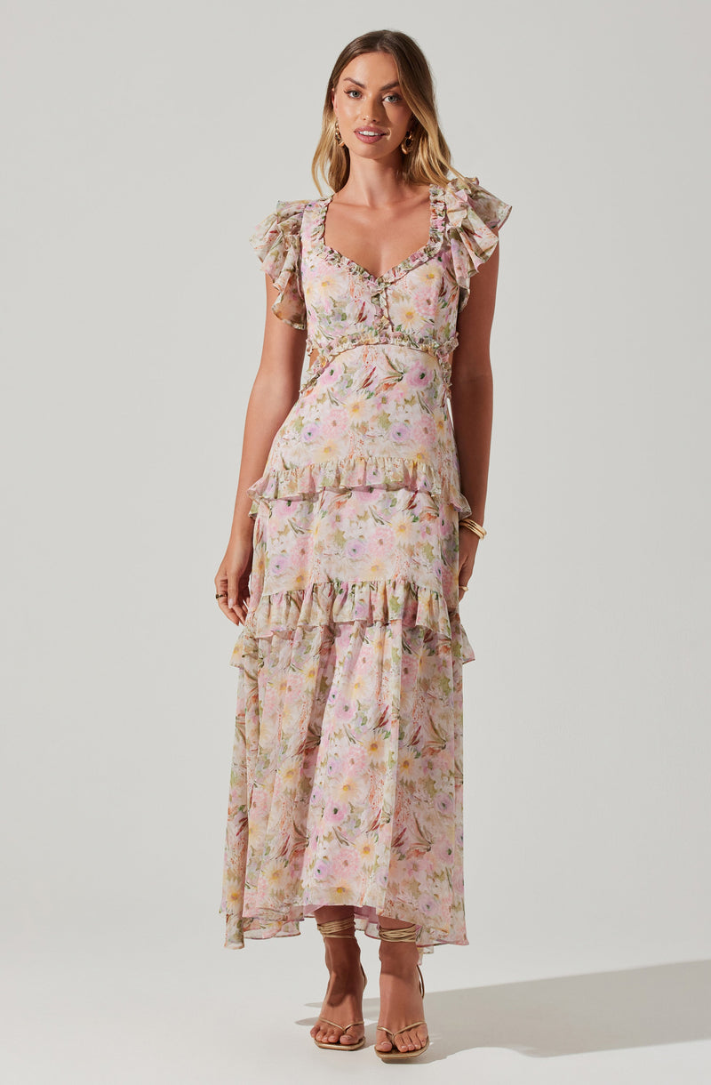 A woman is wearing a long, flowy maxi dress with a pastel floral pattern. The dress features ruffled cap sleeves, a square neckline with a small button detail, and tiered ruffles at the waist and hem. The fabric appears lightweight and breezy, suitable for warm weather. She is also wearing open-toed heels and gold jewelry.