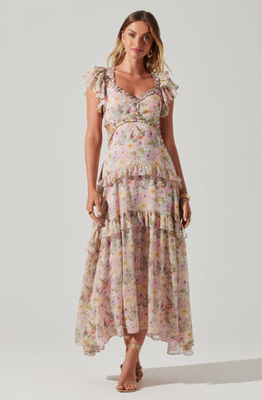 A woman is wearing a flowy, pastel-colored floral dress with a ruffled neckline and cap sleeves. The dress features a fitted bodice with a ruffle around the waist and multiple tiered, ruffled layers in the skirt, creating a soft, romantic look. She is also wearing high-heeled sandals and has gold accessories.