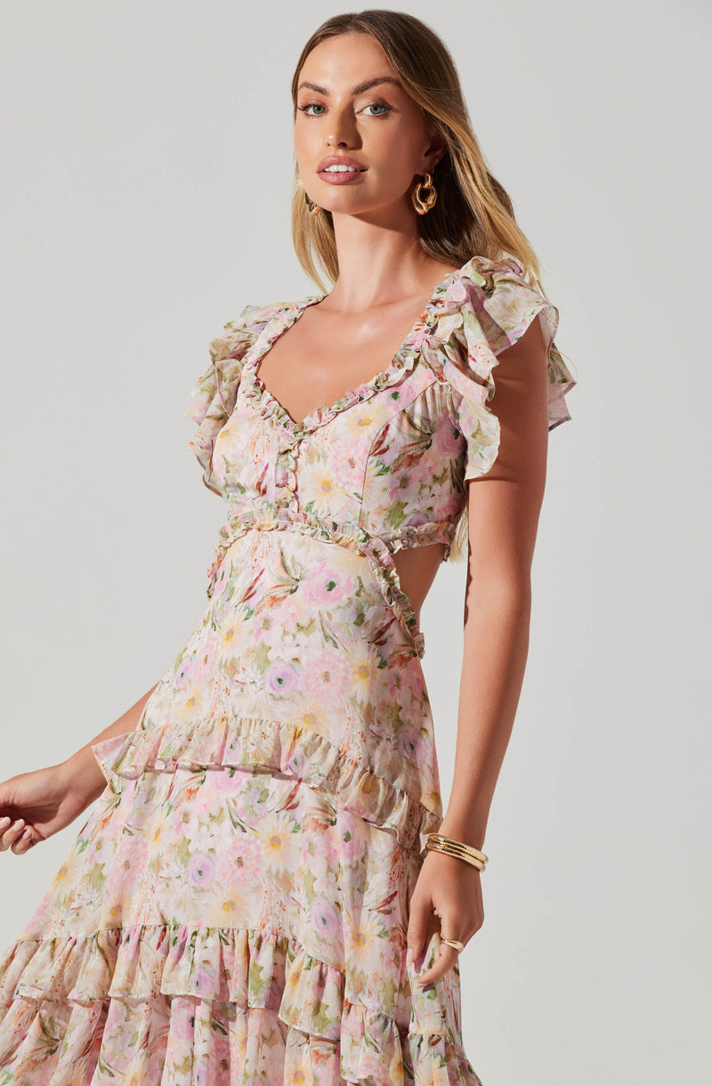 A woman wearing a pastel-colored floral dress with ruffled sleeves and tiered ruffle details. The dress features a fitted bodice with a sweetheart neckline and a flowing skirt with multiple ruffle layers. She accessorizes with gold hoop earrings and gold bangles.