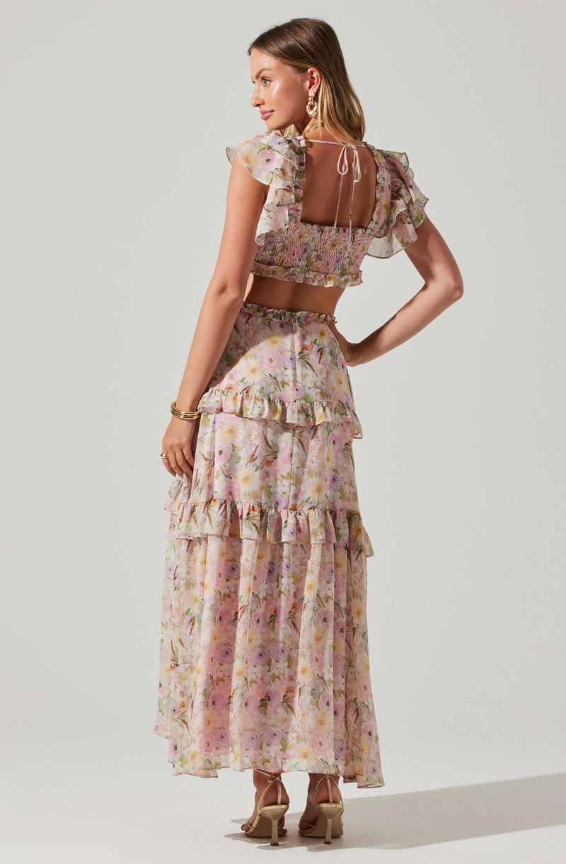 A woman is wearing a two-piece outfit consisting of a cropped top and a long skirt. The top has short, ruffled, semi-sheer sleeves and a smocked bodice, with a square neckline in the back tied with a small bow. The long skirt features a floral pattern with pink, purple, and yellow flowers on a light background, and has ruffled tiers. She is also wearing beige high-heeled sandals, gold hoop earrings, and gold bracelets.