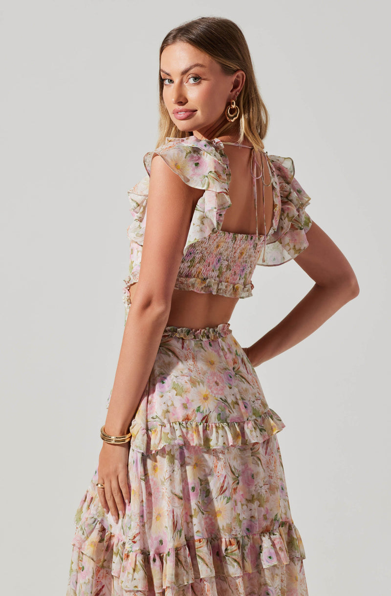 A woman is wearing a two-piece outfit with a floral pattern. The top is a sleeveless, square-neck crop top with ruffled shoulder straps and a smocked, fitted bodice with a tie at the back. The skirt is a high-waisted, ankle-length piece featuring multiple layers of ruffles, all in a pastel-colored floral print.