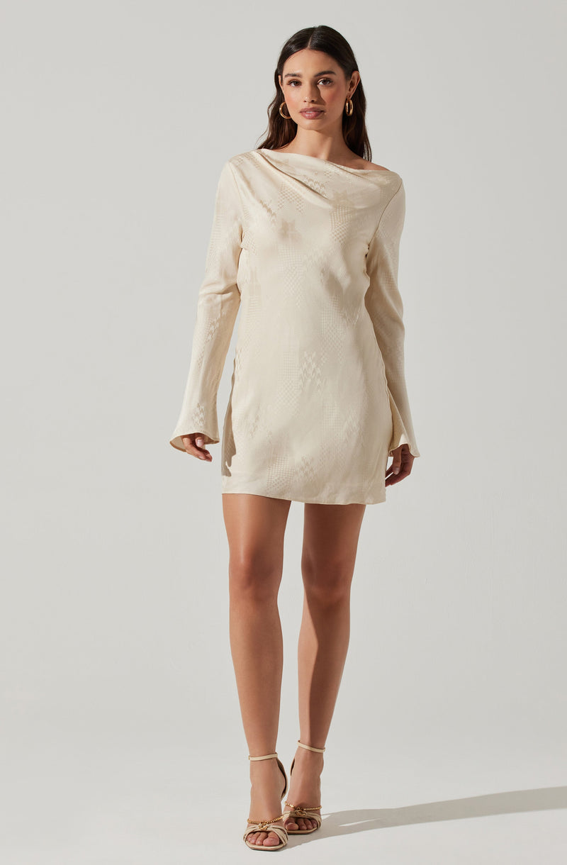 A woman is wearing a cream-colored, long-sleeved mini dress with a subtle textured pattern. The dress features a relaxed fit with a draped neckline that extends over one shoulder. She is also wearing gold hoop earrings and beige strappy high-heeled sandals.