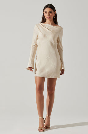 A woman is wearing a cream-colored, long-sleeved mini dress with a subtle textured pattern. The dress features a relaxed fit with a draped neckline that extends over one shoulder. She is also wearing gold hoop earrings and beige strappy high-heeled sandals.