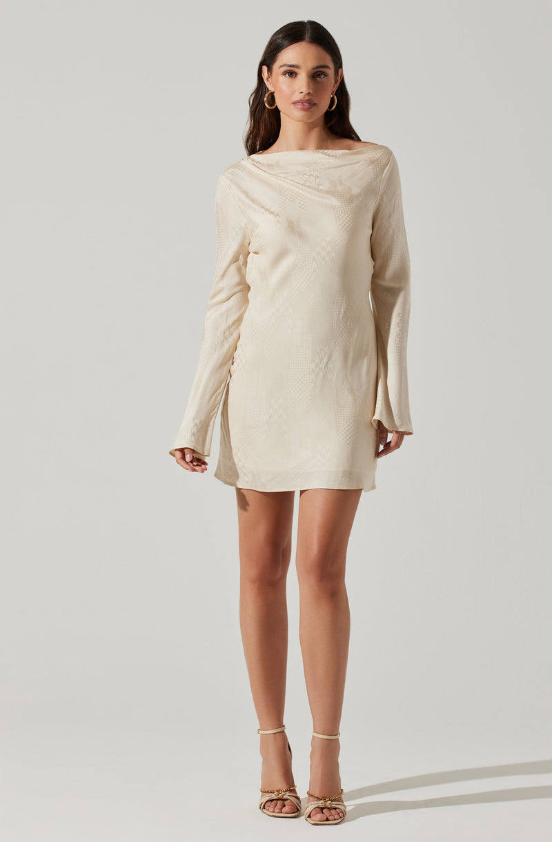 A woman is wearing a cream-colored, long-sleeved dress with a subtle textured pattern. The dress has a loose, relaxed fit with a boat neckline and slightly flared cuffs on the sleeves. She is accessorizing with large hoop earrings and high-heeled sandals with ankle straps.
