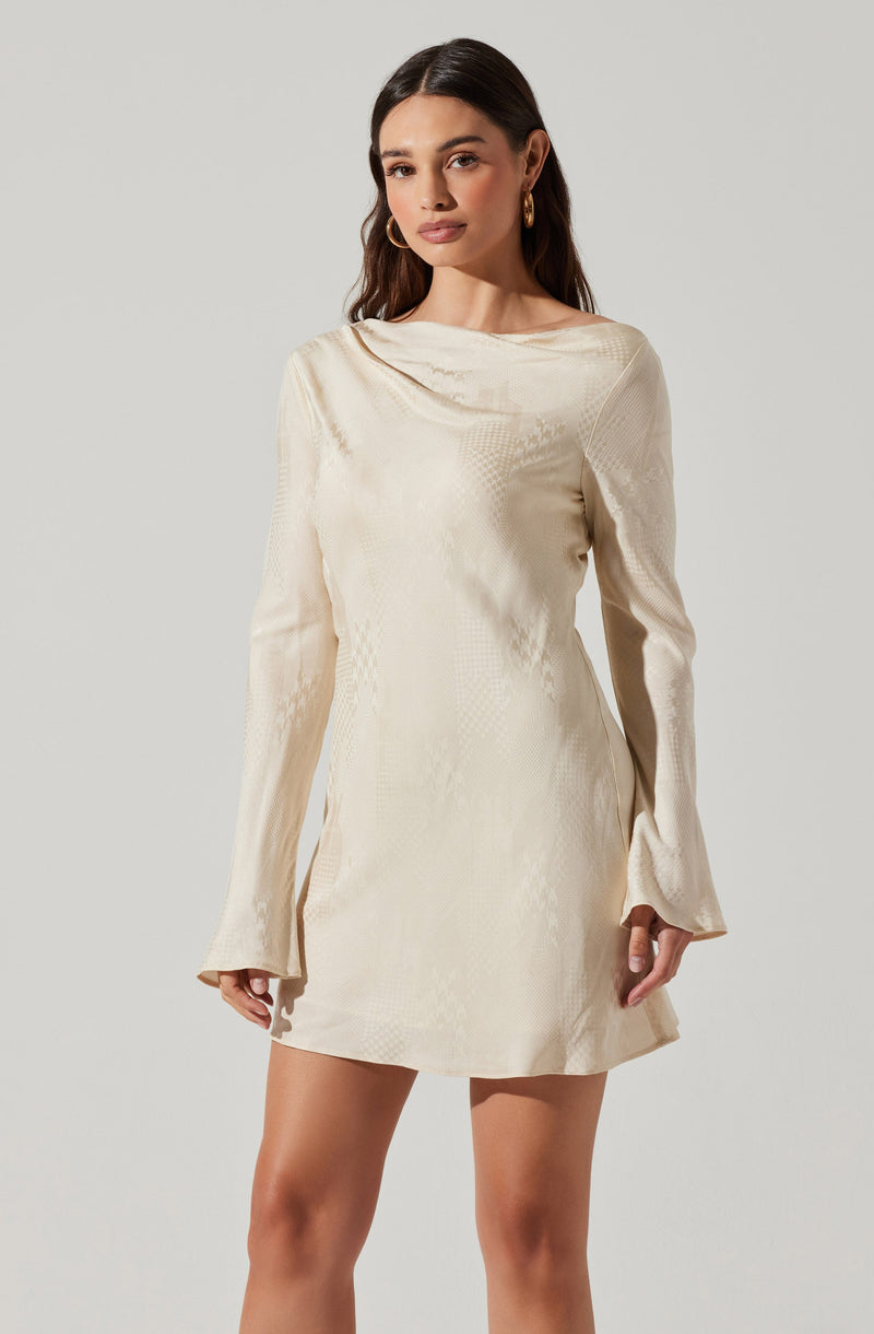 A woman is wearing a cream-colored, long-sleeved mini dress with a subtle shiny, textured pattern. The dress has a high, rounded neckline with a soft, draped collar detail. The sleeves are slightly flared at the cuffs.
