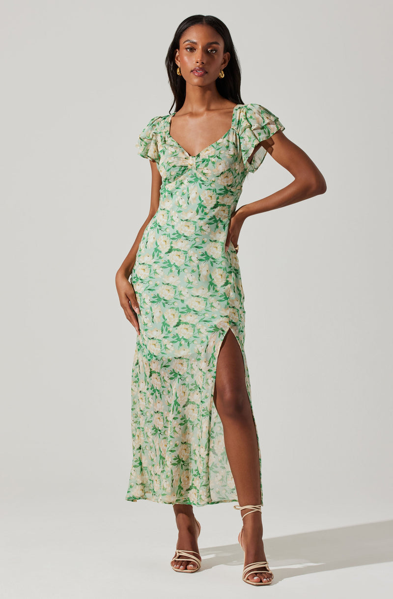 A woman is wearing a long, flowy dress with a light-colored background and a green floral pattern. The dress has short, ruffled sleeves and a V-shaped neckline. It features a high slit on the side, revealing her leg. She is also wearing open-toe high heels and gold earrings.