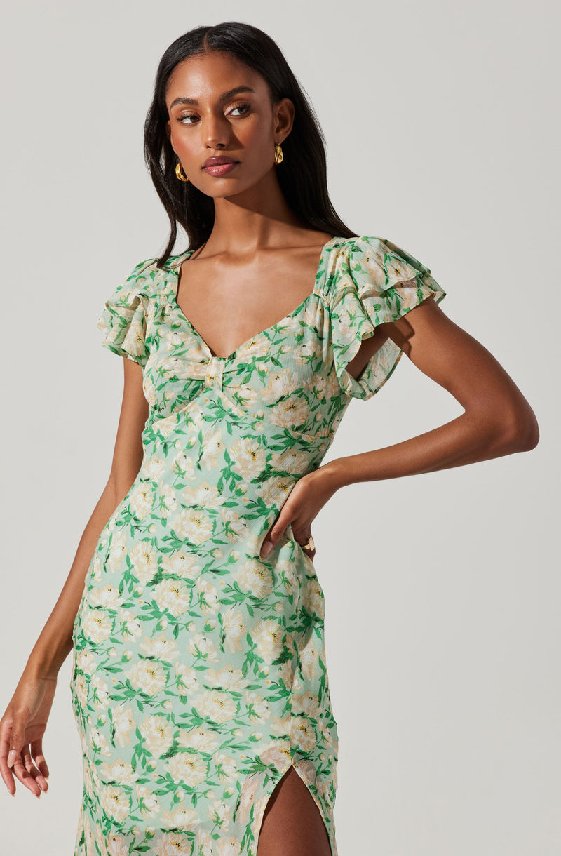 A woman is wearing a light-colored dress with a floral pattern featuring green leaves and white flowers. The dress has a fitted silhouette, a V-neckline, puffed short sleeves, and a front slit on the skirt.