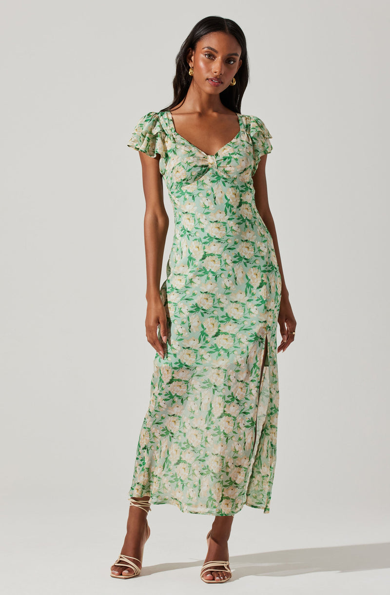 A woman is wearing a long, flowy summer dress with a light cream background and a green floral print. The dress features short, ruffled cap sleeves, a sweetheart neckline with a knotted detail at the center, and a high slit on the side. She is also wearing gold hoop earrings and strappy high-heeled sandals.