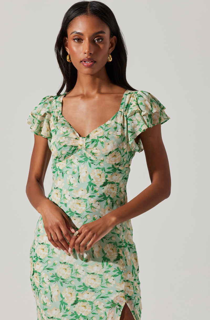 A woman is wearing a light-colored dress with a green floral pattern. The dress features a V-neckline, short ruffled sleeves, and a slit on the side.