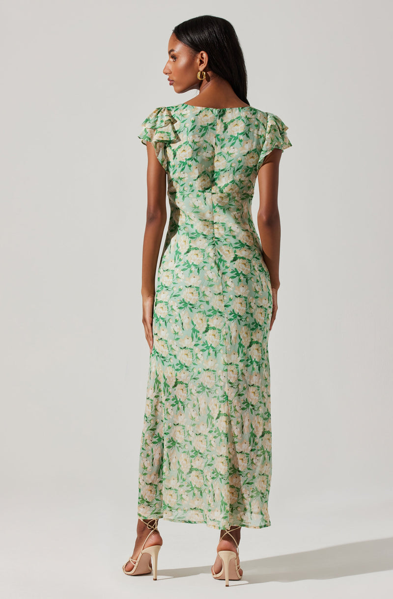 A woman is wearing a long, flowy dress with a floral pattern. The dress has a light green and beige background with darker green leaves and light-colored flowers. It features short, ruffled sleeves and a fitted waist, extending down to her ankles. She is also wearing nude high-heeled sandals.