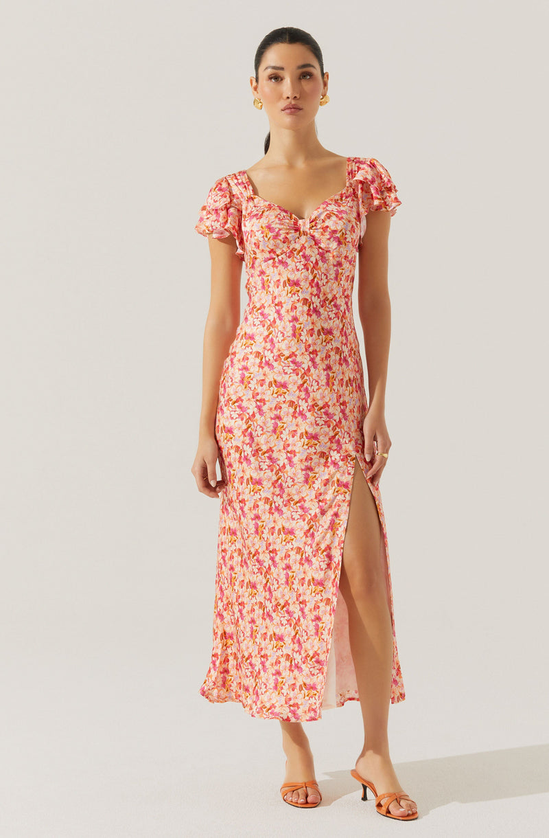 The woman is wearing a long, flowing dress with a pink and orange floral pattern. The dress has short, puffed sleeves and a sweetheart neckline. There is a high slit on the side, revealing her leg. She is also wearing orange high-heeled sandals and gold earrings.