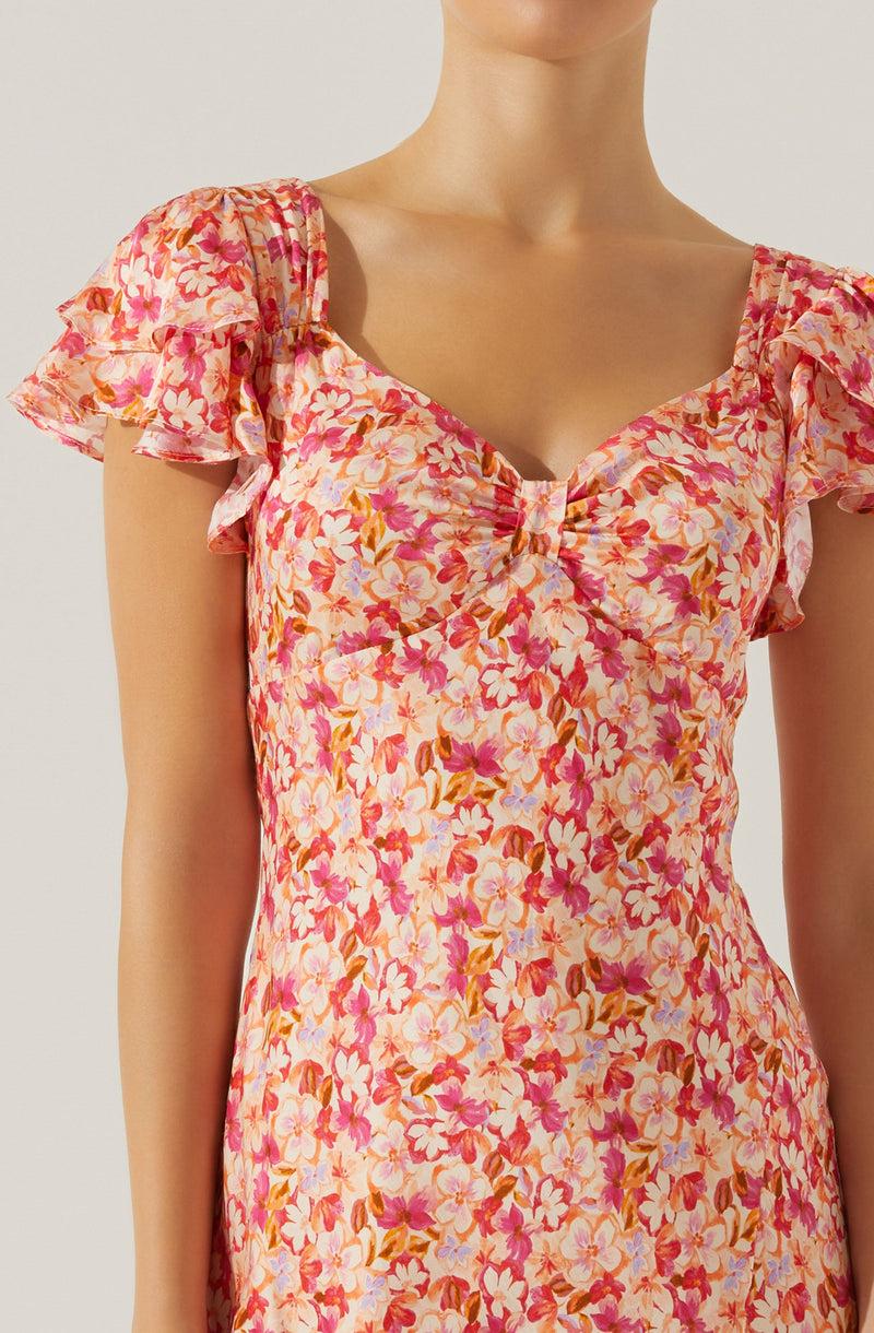 A woman is wearing a dress with a floral pattern featuring pink, white, and light purple flowers with brownish-orange leaves. The dress has a V-shaped neckline with a small pleated detail at the center of the bust. The short sleeves are ruffled and slightly puffed, creating a layered effect.