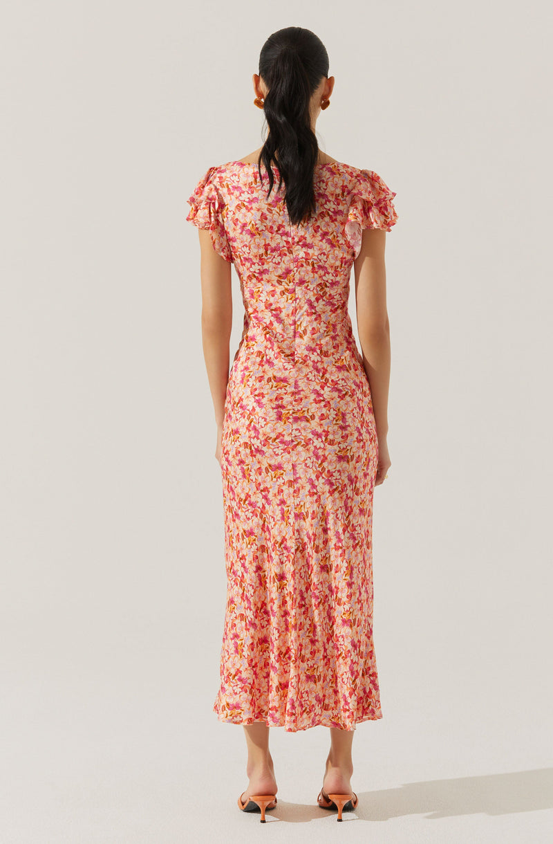 A woman is wearing a long, fitted dress with short, ruffled sleeves. The dress features a colorful, abstract floral pattern in shades of pink, red, orange, and cream. She is also wearing open-toed heels and has her dark hair styled in a ponytail.
