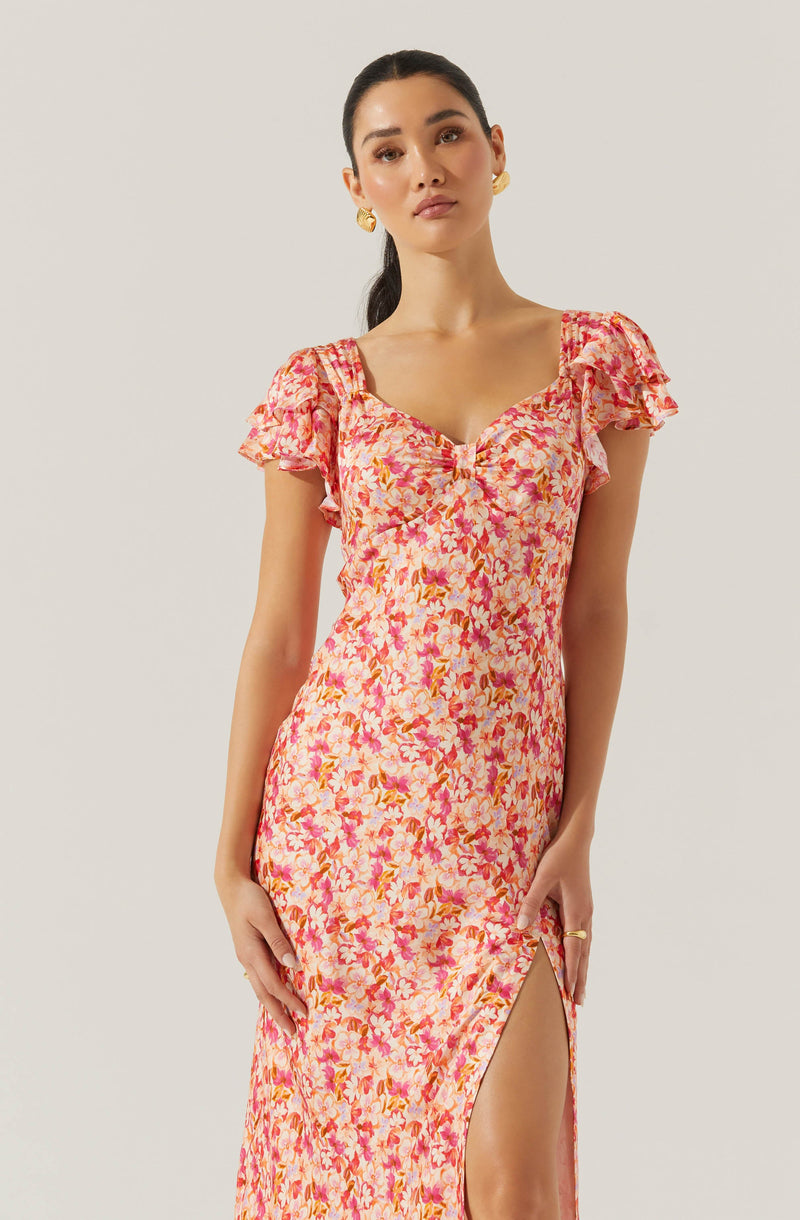 A woman is wearing a long, fitted dress with a pink and orange floral pattern. The dress has short, puffy, ruffled sleeves and a sweetheart neckline. There is a slit on the left side of the dress that reveals part of her leg.