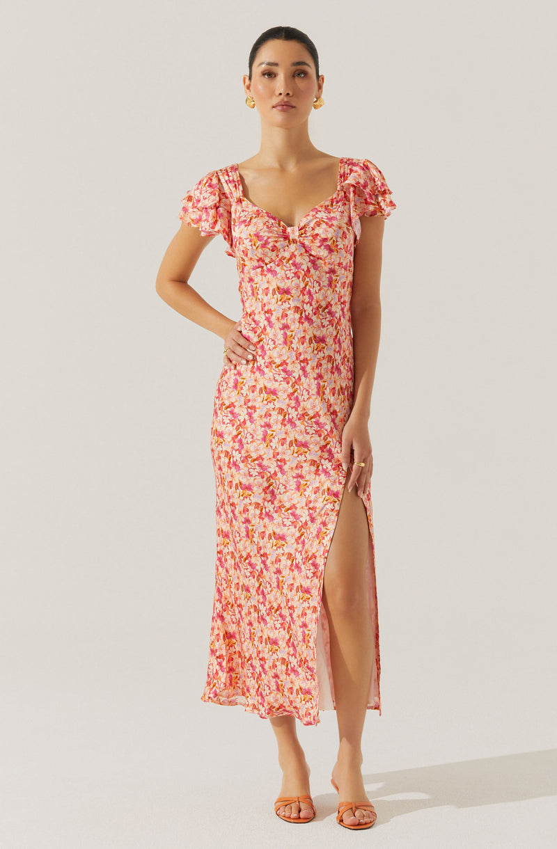 A woman is wearing a long, flowy dress with a pink, orange, and yellow floral pattern. The dress features short, ruffled sleeves, a sweetheart neckline with a small keyhole opening, and a high slit on the left side, revealing part of her leg. She has accessorized with earrings and a ring on her finger. She is wearing open-toe sandals and has her hair pulled back.