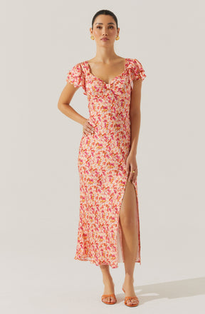 A woman is wearing a long, flowy dress with a pink, orange, and yellow floral pattern. The dress features short, ruffled sleeves, a sweetheart neckline with a small keyhole opening, and a high slit on the left side, revealing part of her leg. She has accessorized with earrings and a ring on her finger. She is wearing open-toe sandals and has her hair pulled back.