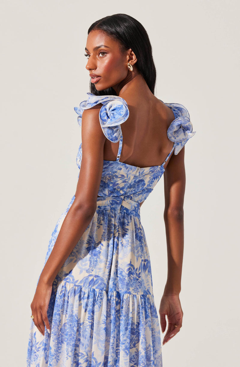 A woman is wearing a sleeveless dress with ruffled shoulder straps and a fitted bodice. The dress features a blue and white floral pattern, with a tiered skirt that has a gathered, flowing silhouette. The fabric appears lightweight and airy.