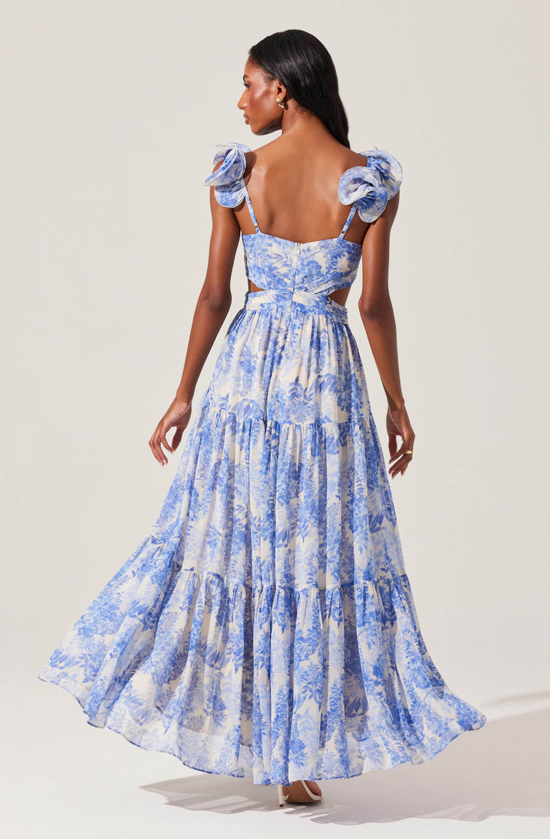A woman is wearing a matching two-piece outfit consisting of a crop top and a long skirt. The crop top has puffy shoulder straps, ruffled details at the shoulders, and a fitted bodice. The skirt is high-waisted, flowing, with a tiered design, and features a blue and white floral pattern.