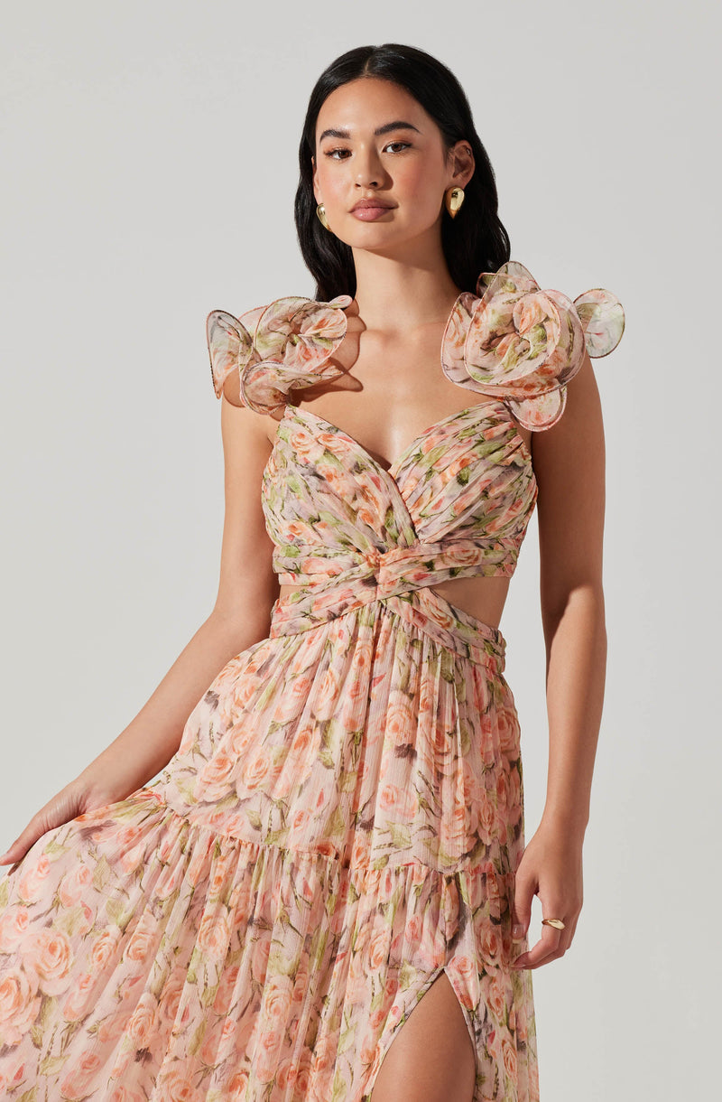 A woman is wearing a flowy, floral dress with a soft pink base and green and peach flower patterns. The dress features dramatic puffed sleeves made from a sheer, ruffled fabric, and a fitted bodice with a twisted detail at the waist. It has a high slit on one side, revealing part of her leg.