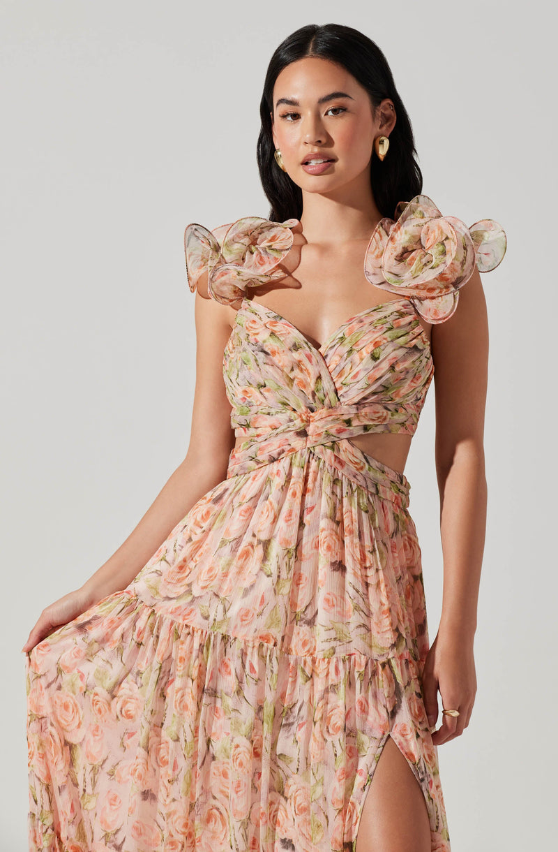 A woman is wearing a dress with a floral pattern of pink, peach, and green roses. The dress features ruffled, puffed sleeves, a sweetheart neckline, and cutouts at the waist. The skirt has a high slit on one side, revealing her leg. The fabric appears lightweight and flowy, with a soft, romantic style.