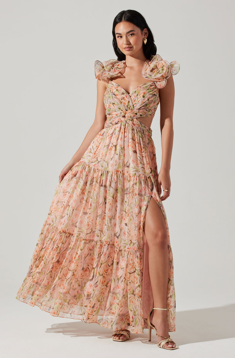 The dress features a floral pattern with soft pink, peach, and green colors. It has puffed, ruffled shoulder details and a deep V-neckline. The bodice is fitted and tied at the waist with a knot, creating a cutout effect at the midsection. The skirt is long, flowing, and tiered with a high slit on one side, revealing the leg and a high-heeled sandal.