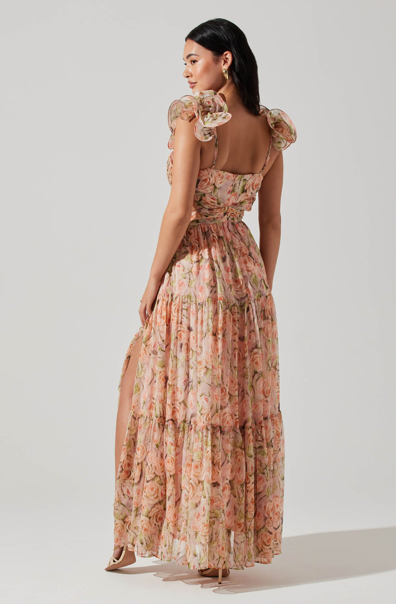 A woman is wearing a long, flowy dress with a floral pattern in shades of pink, peach, and green. The dress features ruffled, puffed sleeves made from sheer fabric, and it has a fitted bodice with thin spaghetti straps. The skirt is tiered and reaches down to the floor, with a slit on one side revealing her leg. She is also wearing high-heeled shoes and gold earrings.