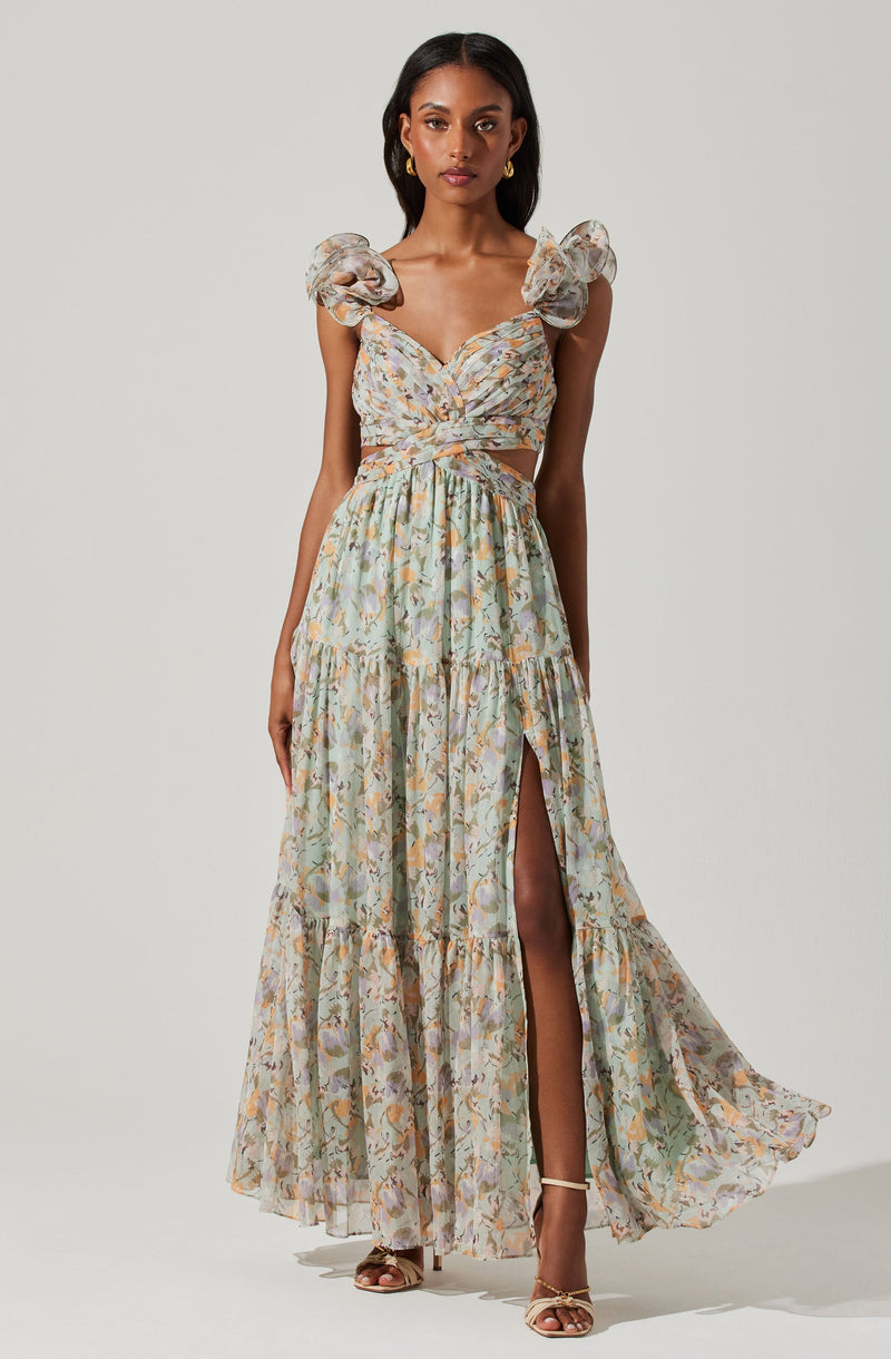 A woman is wearing a long, flowy dress with a floral pattern in soft pastel shades. The dress features puffed, ruffled sleeves, a fitted bodice with a twist detail at the front, and a high slit on one side, revealing her leg. She is also wearing strappy heels.
