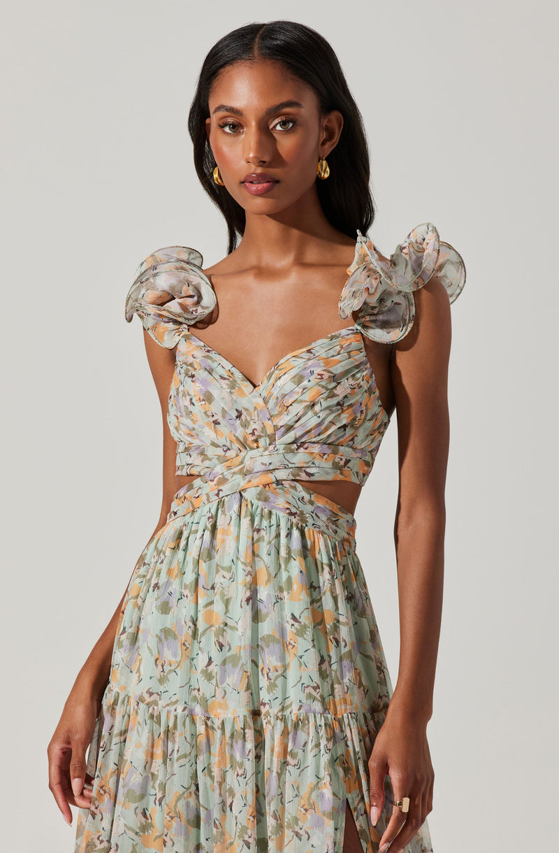 A young woman is wearing a sleeveless, cropped dress with ruffled shoulder details. The fabric has a soft, pastel-colored floral pattern with shades of light green, peach, beige, and purple. The top of the dress features a sweetheart neckline with ruched detailing, and the skirt is high-waisted and flowy, reaching down to a midi length with a slit on one side.
