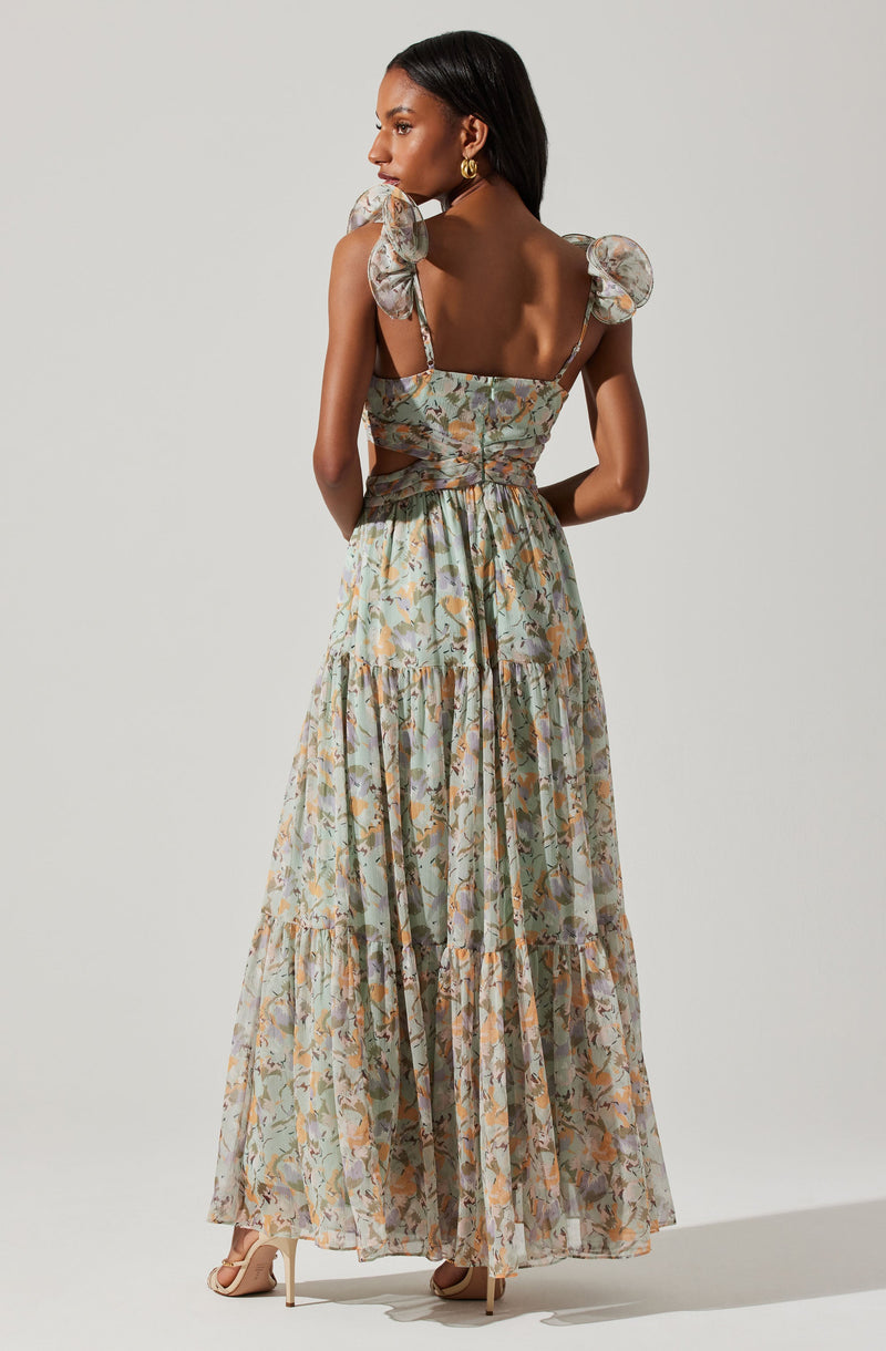 The dress is a sleeveless, maxi-length style with a floral pattern in soft pastel colors. It has puffed shoulder straps tied into bows and a fitted bodice with cutout details on the sides. The fabric appears lightweight and flowy, creating a romantic and elegant look.