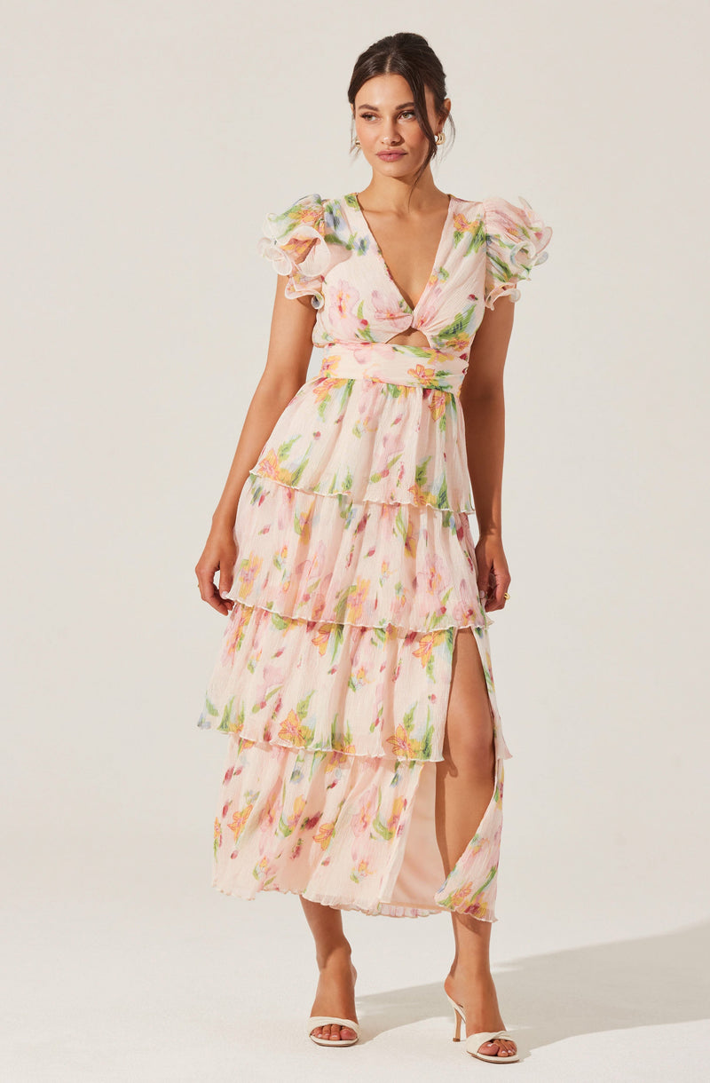 A woman is wearing a long, flowing dress with a pastel floral pattern. The dress features short, puffed sleeves with ruffled edges, a V-neckline, and a fitted waist. It has multiple layered ruffles descending to the hem, with a slit on the left side revealing her leg. She is also wearing white high-heeled sandals.