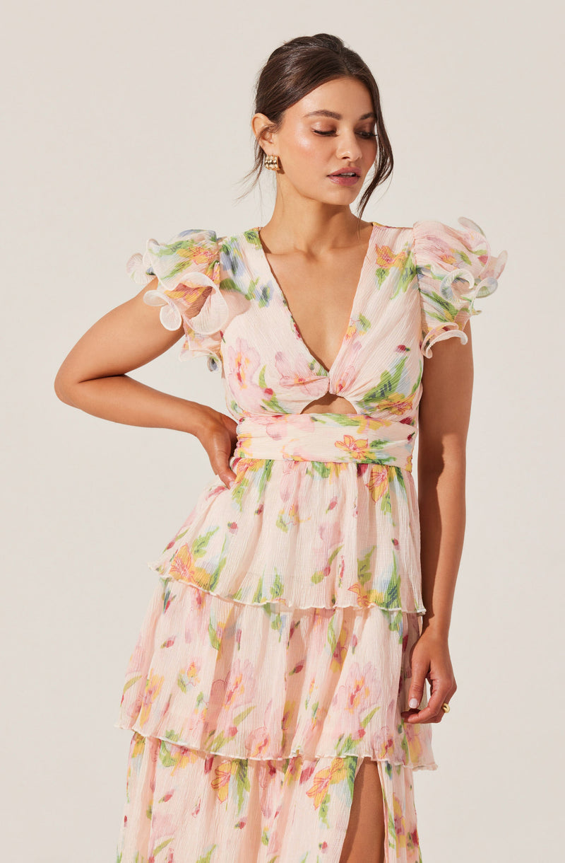 A woman is wearing a light-colored, floral dress with soft pastel shades of pink, green, and yellow. The dress features ruffled cap sleeves, a V-neckline with a twist knot detail at the bust, and a tiered skirt with a slit at the front. The fabric appears to be lightweight and textured.