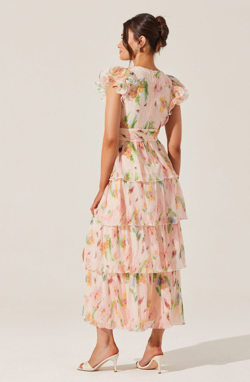 A woman is wearing a long, flowy dress with a pastel floral pattern. The dress features a high waistline with a matching fabric belt, and the fabric is lightweight and semi-sheer. The dress has short, ruffled sleeves with layered, tiered skirts that create a tiered effect from the waist down. She is also wearing high-heeled shoes in a neutral color.