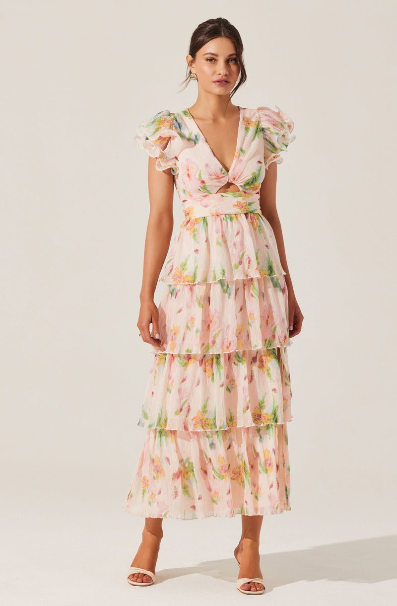 A woman is wearing a long, tiered dress with a floral pattern featuring pink, yellow, and green colors. The dress has a deep V-neckline with a twisted knot detail at the bust, short puff sleeves.